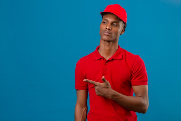 Young african american delivery man wearing red polo shirt and cap à l'écart et pointant le doigt sur le côté debout sur bleu isolé