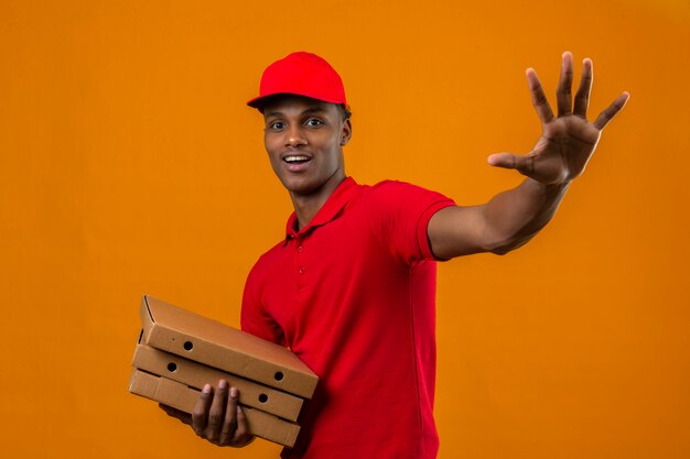 Young african american delivery man wearing red polo shirt and cap holding package saluting with hand with happy expression over isolated orange