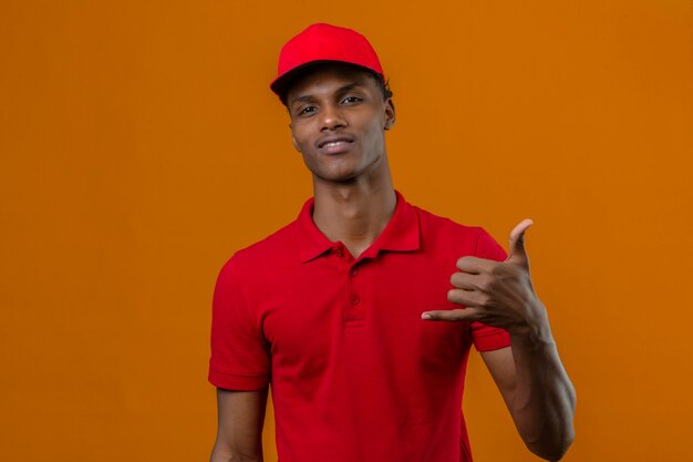 Young african american delivery man wearing red polo shirt and cap looking confiant making phone geste call me back sign over isolated orange