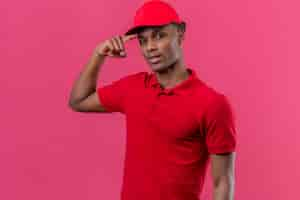 Photo gratuite young african american delivery man wearing red polo shirt and cap with smile on face pointant to head with finger over isolated pink