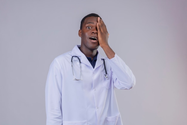 Young African American male doctor wearing white coat avec stéthoscope couvrant un œil avec la main à la surprise et heureux