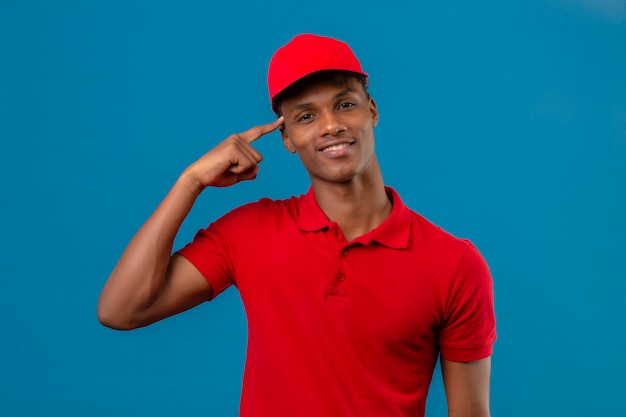 Young african american smart à la livraison homme portant un polo et une casquette rouge souriant et pointant sur sa tête debout sur bleu isolé