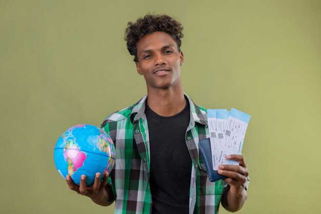 Young african american traveller man holding globe et billets d'avion regardant la caméra en souriant debout sur fond vert