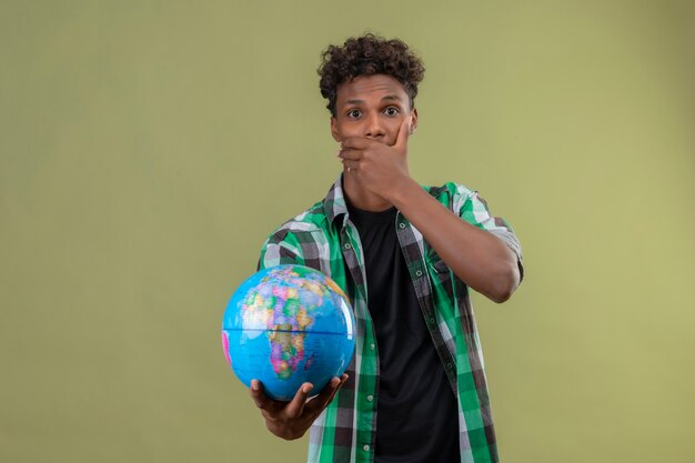 Young african american traveller man holding globe looking at camera choqué couvrant la bouche avec la main debout sur fond vert