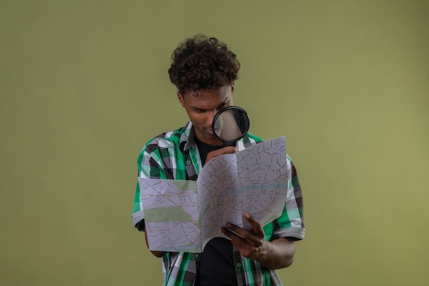 Photo gratuite young african american traveller man holding map en le regardant à travers une loupe debout sur fond vert