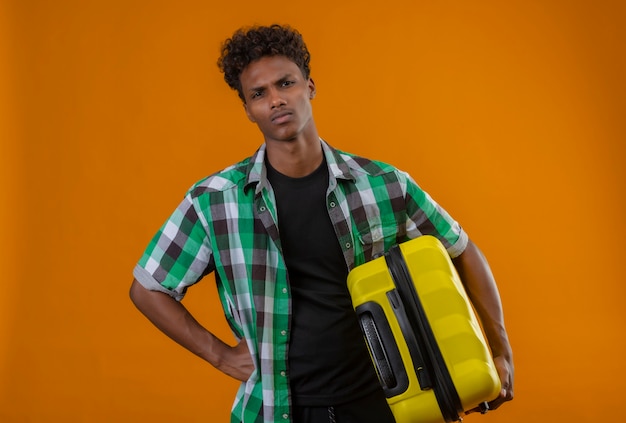 Young african american traveller man holding valise regardant la caméra mécontent de froncer les sourcils debout sur fond orange