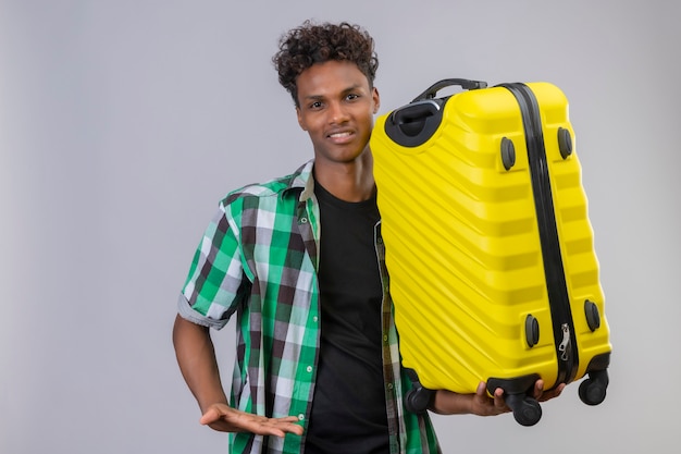 Young african american traveller man holding valise regardant la caméra souriant joyeusement positif et heureux debout sur fond blanc