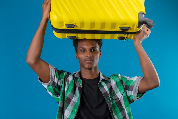 Photo gratuite young african american traveller man holding valise sur sa tête regardant la caméra avec une expression sérieuse confiante sur le visage debout sur fond bleu