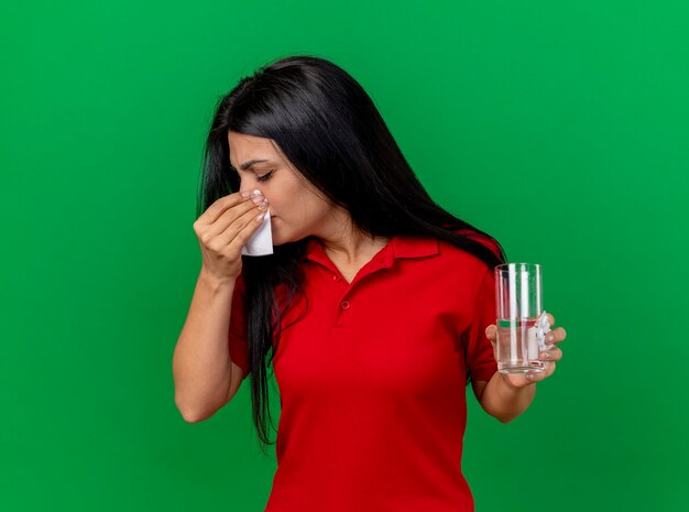 Young caucasian malade girl holding pack de comprimés verre d'eau et essuyant le nez avec une serviette avec les yeux fermés isolé sur fond vert avec copie espace