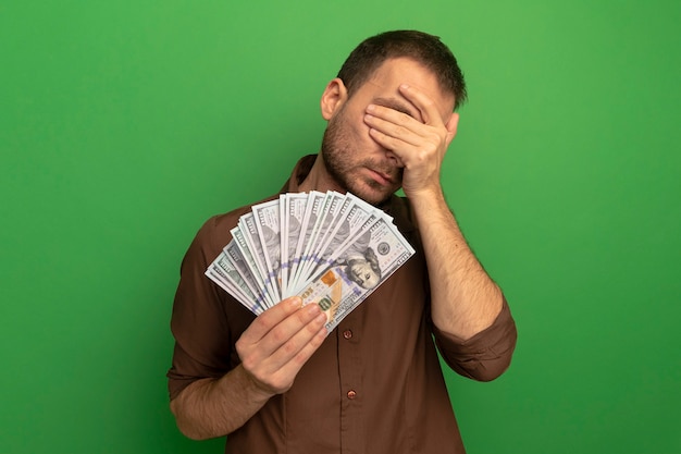 Photo gratuite young caucasian man holding money couvrant les yeux avec la main isolé sur fond vert avec copie espace