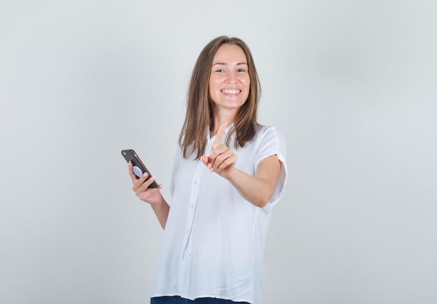 Young female holding smartphone et gestes non avec le doigt en chemise blanche