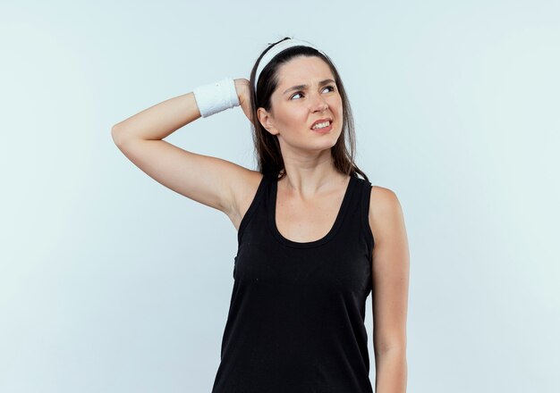 Young fitness woman in headband à côté avec la main sur la tête pour erreur debout sur un mur blanc