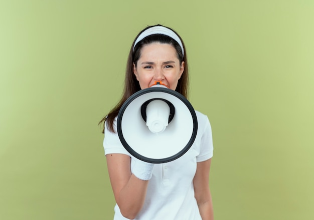 Young fitness woman in headband criant au mégaphone bruyamment debout sur fond clair