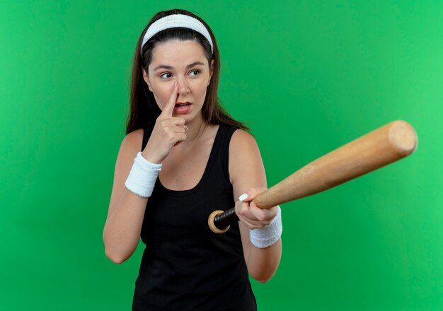 Young fitness woman in headband holding baseball bat à côté en pointant avec le doigt ot son nez debout sur fond vert