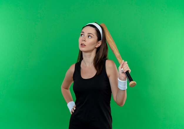 Young fitness woman in headband holding baseball bat loking de côté avec expression pensive debout sur fond vert