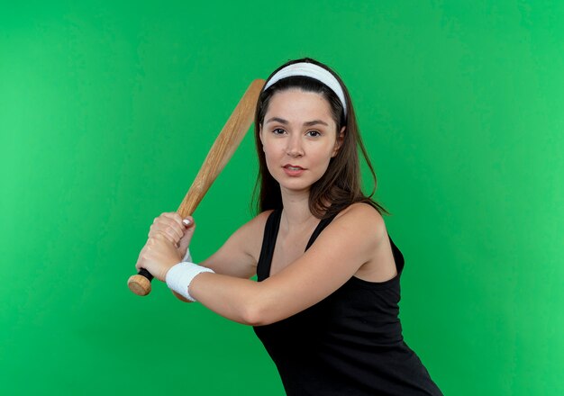 Young fitness woman in headband swinging baseball bat à confiant debout sur mur vert