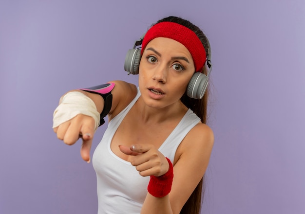 Photo gratuite young fitness woman in sportswear avec bandeau et casque pointant avec le doigt à la caméra inquiète debout sur mur gris