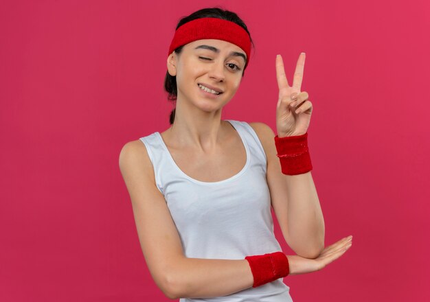 Young fitness woman in sportswear avec bandeau clignotant et souriant montrant le signe de la victoire debout sur le mur rose