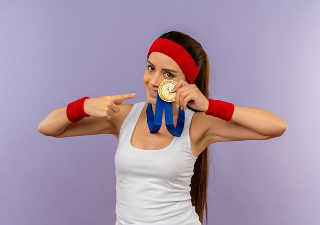 Young fitness woman in sportswear avec bandeau avec médaille d'or autour de son cou montrant et pointant avec le doigt sur elle souriant debout sur mur gris