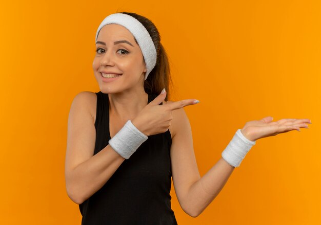 Young fitness woman in sportswear avec bandeau pointant heureux et positif avec les bras et le doigt sur le côté debout sur le mur orange