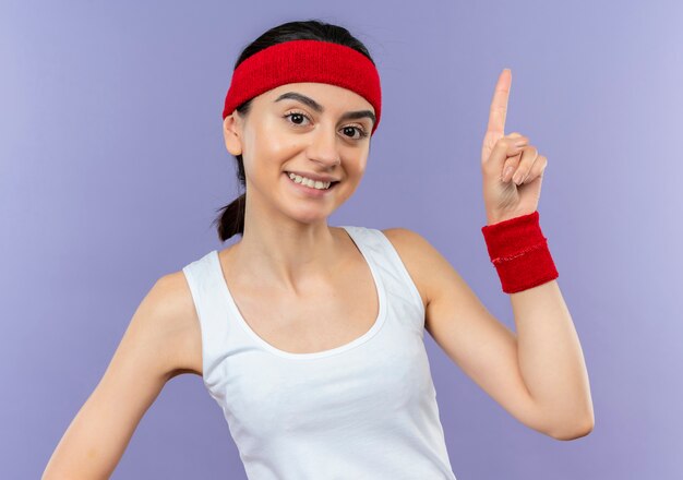 Young fitness woman in sportswear avec bandeau pointant vers le haut avec l'index souriant confiant debout sur mur violet