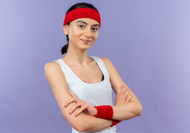 Young fitness woman in sportswear avec bandeau avec sourire confiant avec les mains croisées sur la poitrine debout sur le mur violet