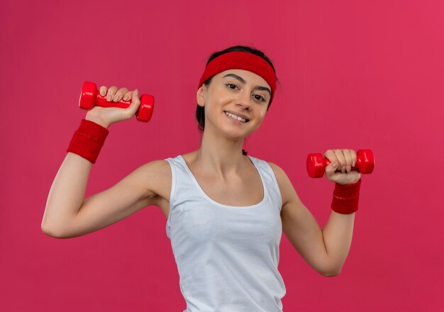 Young fitness woman in sportswear avec bandeau tenant deux haltères dans les mains levées souriant confiant faisant des exercices debout sur un mur rose