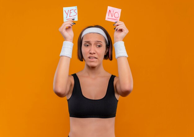 Young fitness woman in sportswear avec bandeau tenant deux papiers de rappel avec mot oui et non dans les mains levées à la confusion debout sur le mur orange