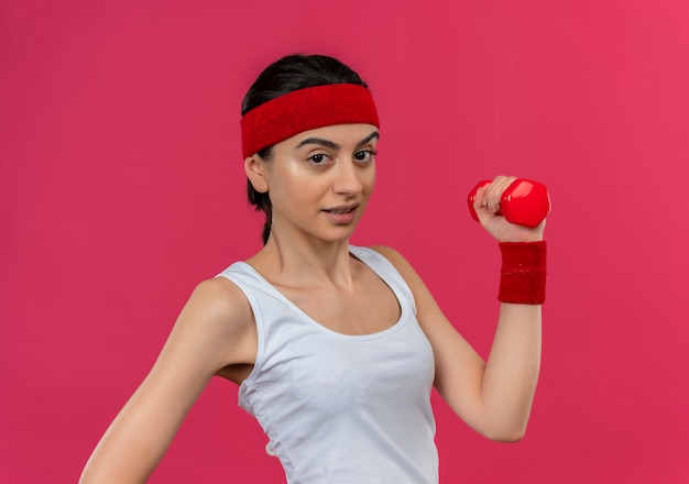 Young fitness woman in sportswear avec bandeau tenant haltère dans la main levée à la confiance en faisant des exercices debout sur un mur rose
