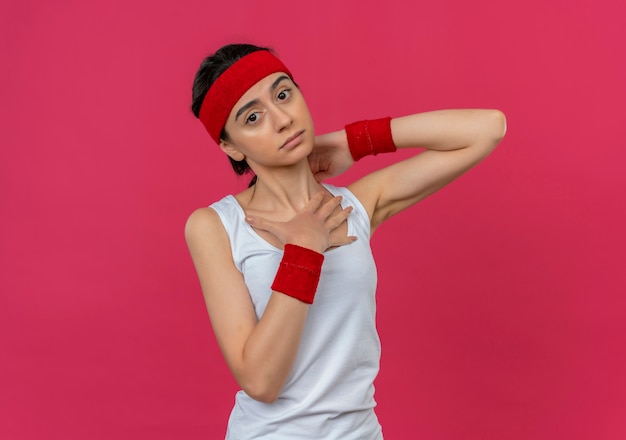 Young fitness woman in sportswear avec bandeau touchant son cou mécontent de se sentir mal à l'aise debout sur un mur rose