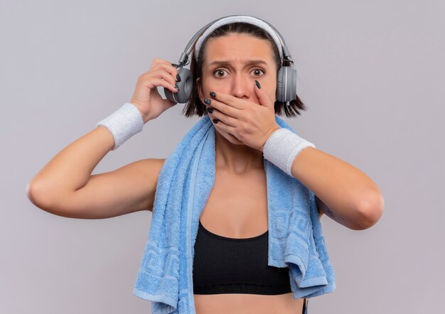 Young fitness woman in sportswear avec un casque sur la tête et une serviette sur son cou choqué couvrant la bouche avec la main debout sur un mur blanc