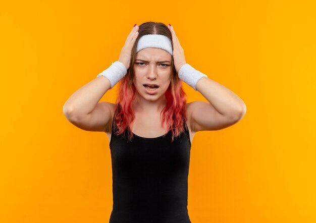 Young fitness woman in sportswear à la confusion et très déçu de toucher sa tête debout sur un mur orange