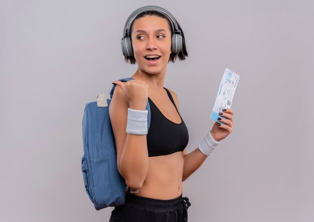 Young fitness woman in sportswear avec des écouteurs sur la tête avec sac à dos tenant billet d'avion à côté pointant vers l'arrière avec le pouce debout sur un mur blanc