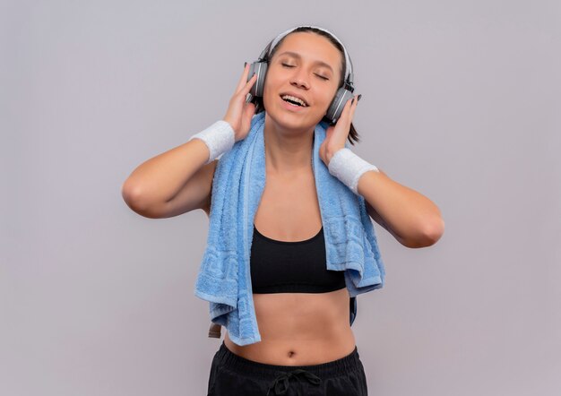 Young fitness woman in sportswear avec des écouteurs sur la tête et une serviette sur son cou appréciant sa musique préférée avec les yeux fermés debout sur un mur blanc