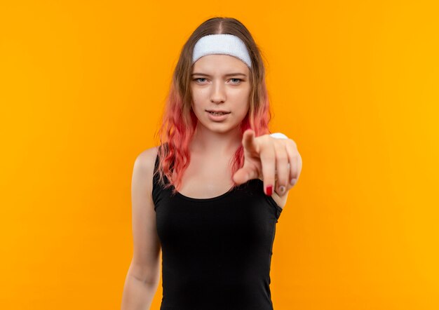 Young fitness woman in sportswear pointant avec l'index à la caméra avec un visage sérieux debout sur un mur orange