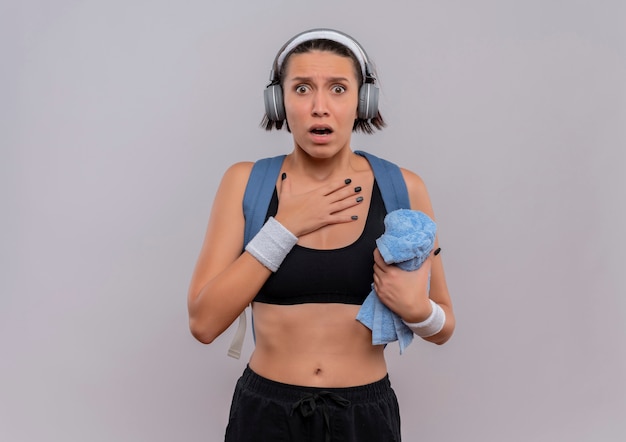 Young fitness woman in sportswear avec sac à dos et casque sur la tête tenant une serviette avec la main sur sa poitrine peur debout sur un mur blanc