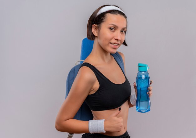 Young fitness woman in sportswear avec sac à dos et tapis de yoga tenant une bouteille d'eau souriant montrant les pouces vers le haut debout sur un mur blanc