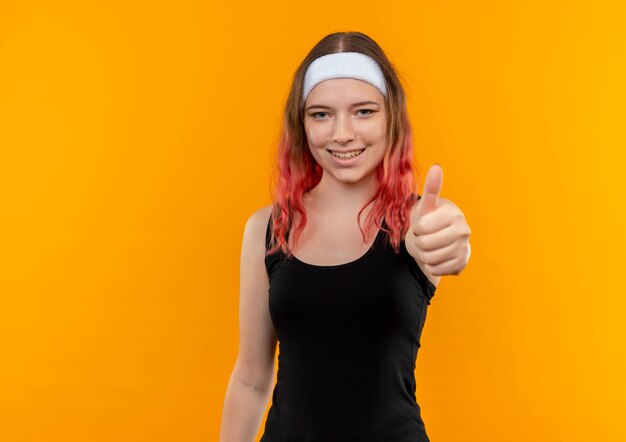 Young fitness woman in sportswear souriant gaiement montrant les pouces vers le haut debout sur le mur orange