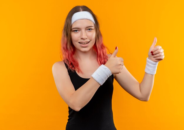 Young fitness woman in sportswear souriant gaiement montrant les pouces vers le haut avec les deux mains debout sur le mur orange