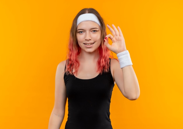 Young fitness woman in sportswear souriant joyeusement montrant signe ok debout sur un mur orange