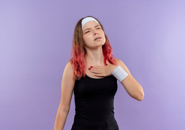 Young fitness woman in sportswear tenant la main sur sa poitrine à la fatigue debout sur le mur violet