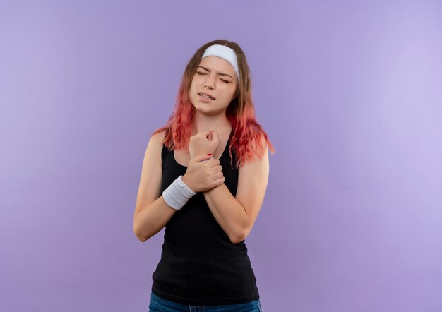 Young fitness woman in sportswear touchant sa main ressentant de la douleur debout sur le mur violet