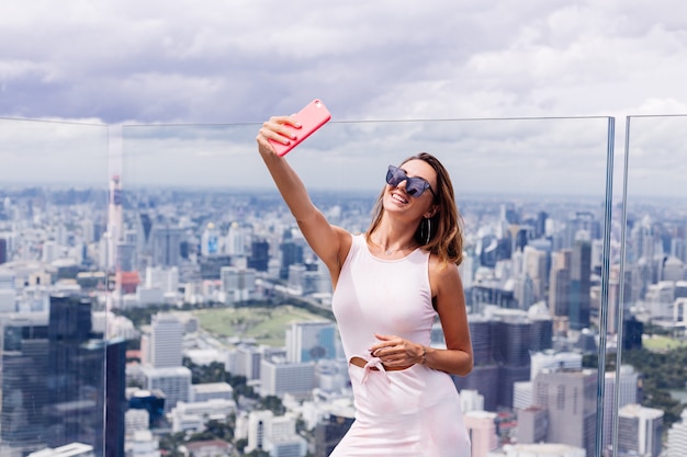 Young happy smiling caucasian woman traveller en robe de montage et lunettes de soleil à l'étage élevé à Bangkok holding phone