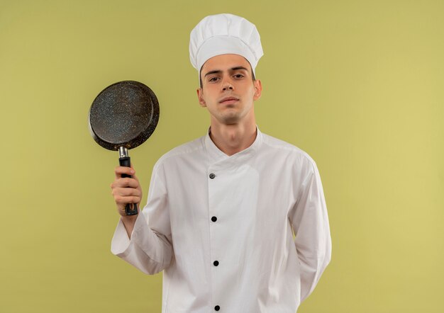 Young male cook wearing chef uniform holding poêle à frire sur mur vert isolé avec copie espace