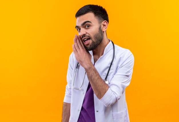 Young male doctor wearing stéthoscope blouse médicale chuchote sur mur jaune isolé