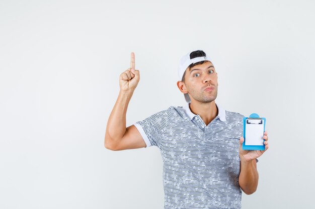 Young male holding clipboard, pointant vers le haut en t-shirt et casquette et à la recherche concentrée