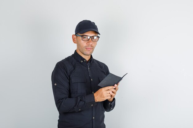 Young man holding mini notebook en chemise noire et casquette