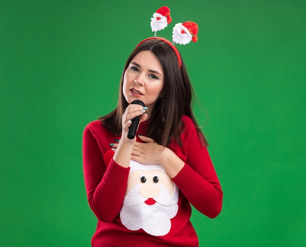 Young pretty caucasian girl wearing Santa Claus pull et bandeau parler dans le microphone en gardant la main sur la poitrine isolée sur le mur vert avec copie espace