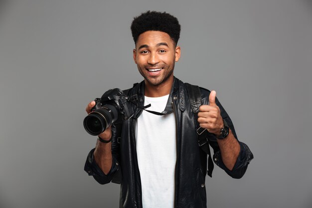 Young smiling afro american man holding photocamera et montrant le pouce vers le haut de geste, à la recherche