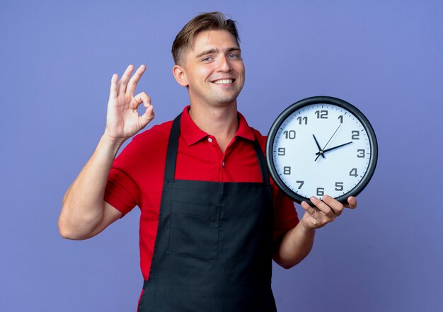 Young smiling blonde male barber en uniforme tient horloge gesticulant signe de la main ok isolé sur l'espace violet avec copie espace